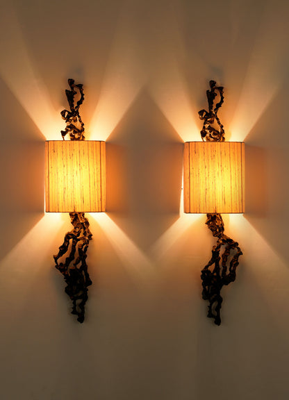 The VIYA Andaman Wall Sconce A features a square beige fabric shade that emits warm light with starburst patterns, while brass details and decorative dark metal elements evoke the organic forms of the Andaman Islands.