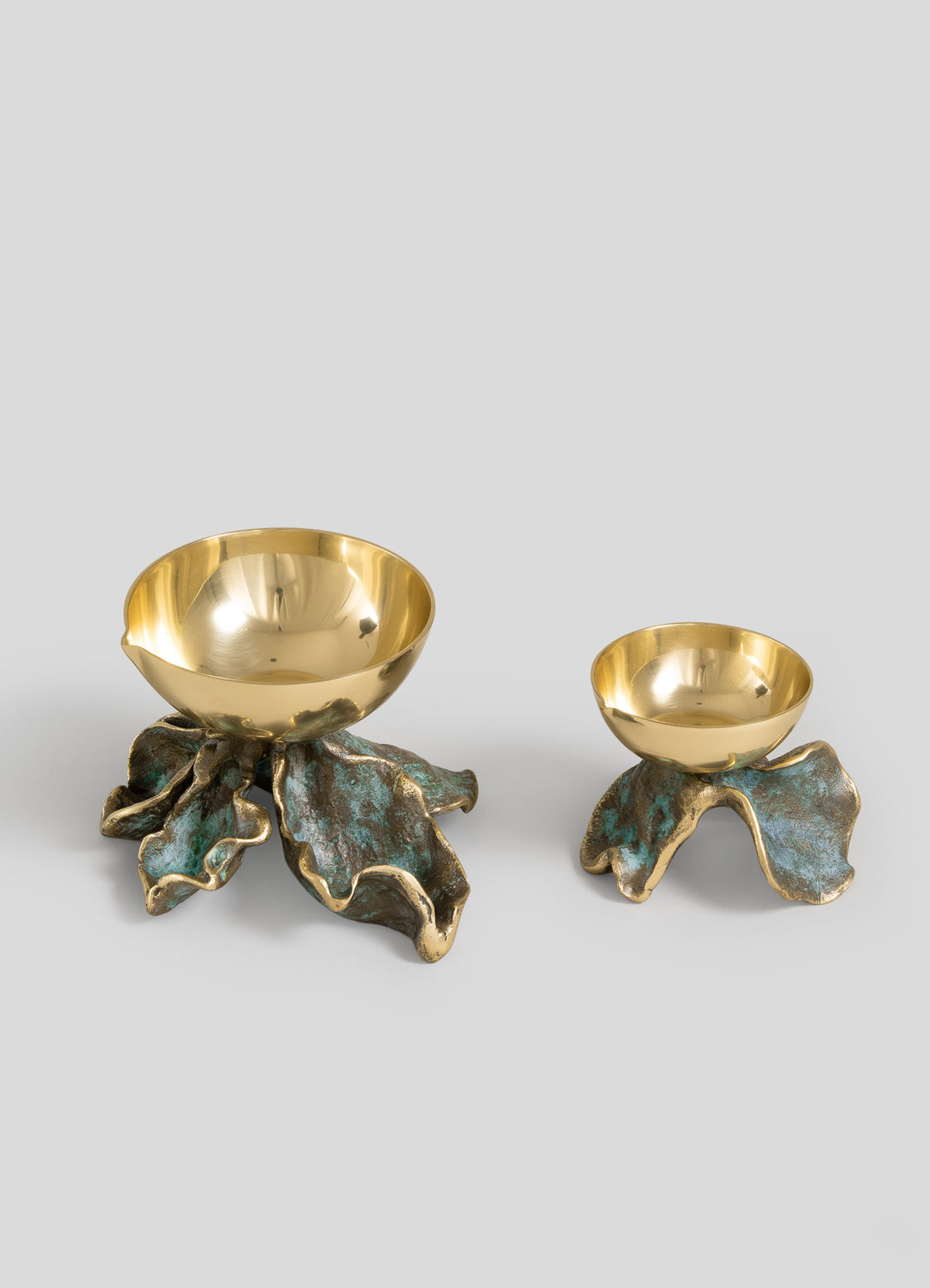 Two gold-colored bowls from Viya, each supported by sculpted, patinaed leaf-shaped bases reminiscent of botanical candle holders, are displayed on a plain gray background. The larger bowl alongside the Rain Song Diya Small adds an elegant touch, perfect for Indian monsoon decor.