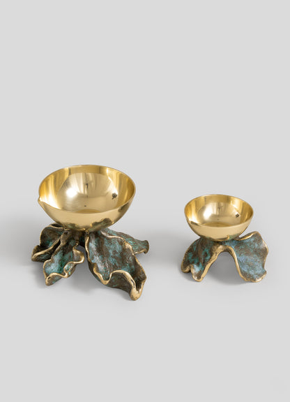 Two gold-colored bowls from Viya, each supported by sculpted, patinaed leaf-shaped bases reminiscent of botanical candle holders, are displayed on a plain gray background. The larger bowl alongside the Rain Song Diya Small adds an elegant touch, perfect for Indian monsoon decor.