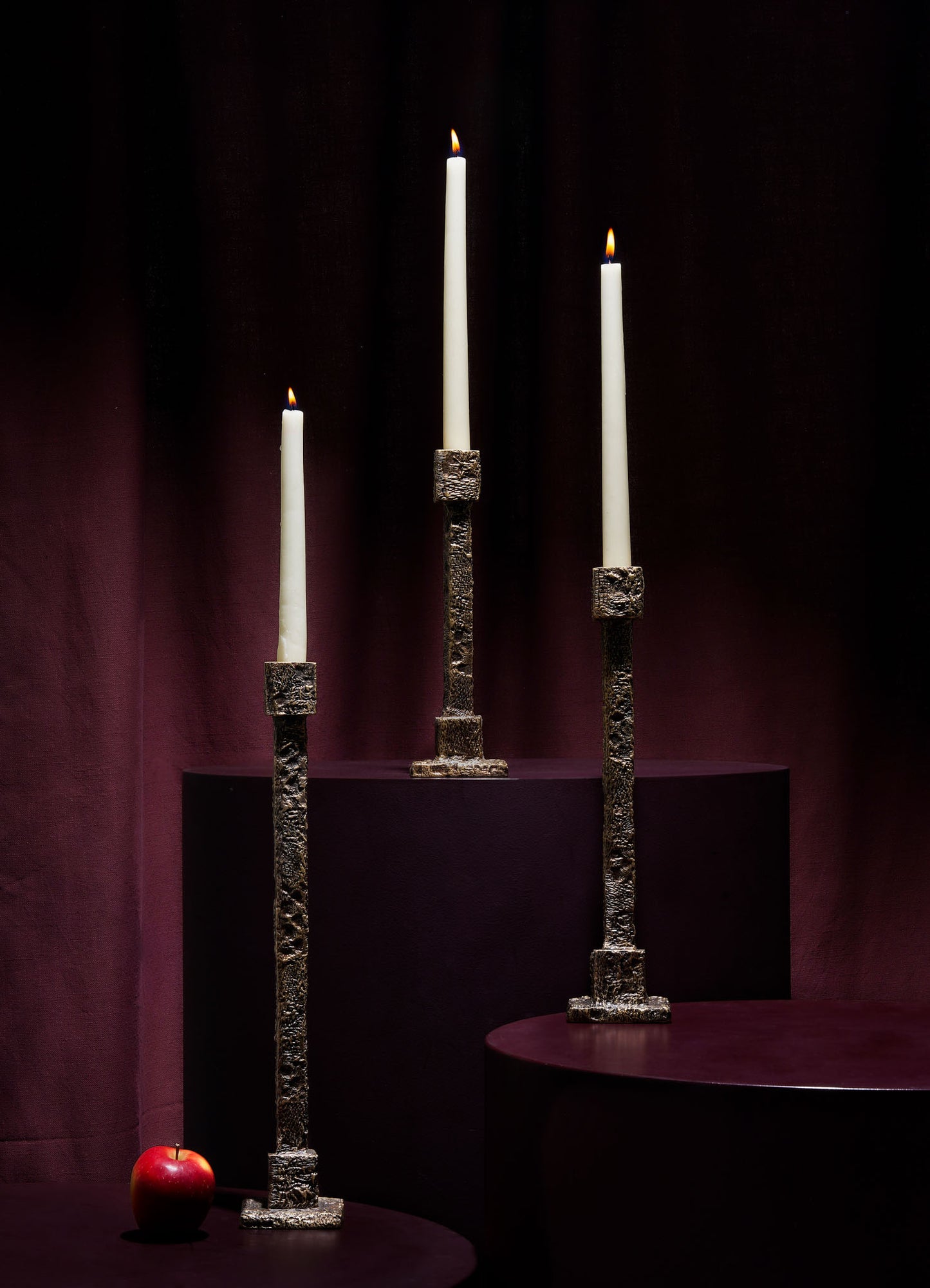 Three lit white candles in VIYA Tiruppur Candle Holder Mediums stand on burgundy pedestals against a dark backdrop. A red apple rests on the lowest pedestal, enhancing the rich, brocade-like ambiance of the arrangement.