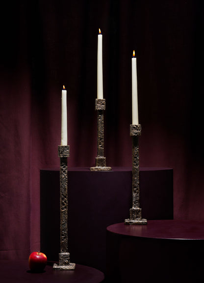 Three lit white candles in VIYA Tiruppur Candle Holder Mediums stand on burgundy pedestals against a dark backdrop. A red apple rests on the lowest pedestal, enhancing the rich, brocade-like ambiance of the arrangement.