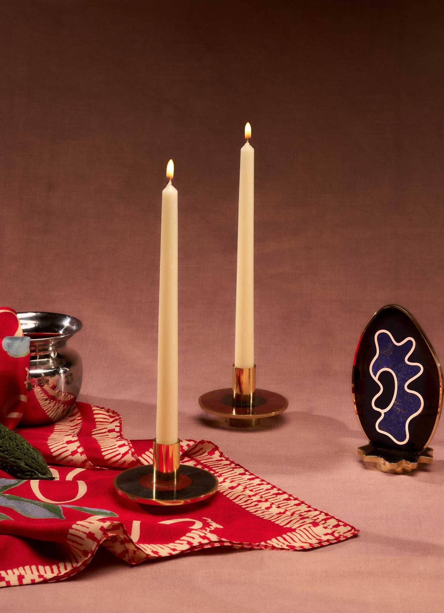 Two cream-colored taper candles in VIYA Alta Candle Holders rest on a red patterned cloth inspired by Indian Alta dye, next to a silver bowl and an abstract egg-shaped blue, white, and black art piece on a gold stand.