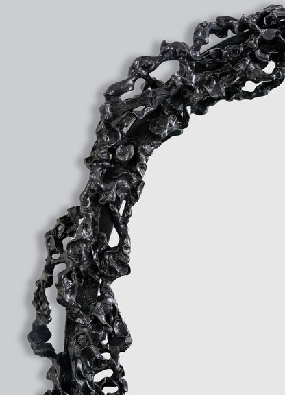 Close-up of the Andaman Mirror Large by VIYA—a black sculptural frame with an irregular, organic texture resembling twisted metal—curving and partially enclosing a blank white space against a plain gray background.