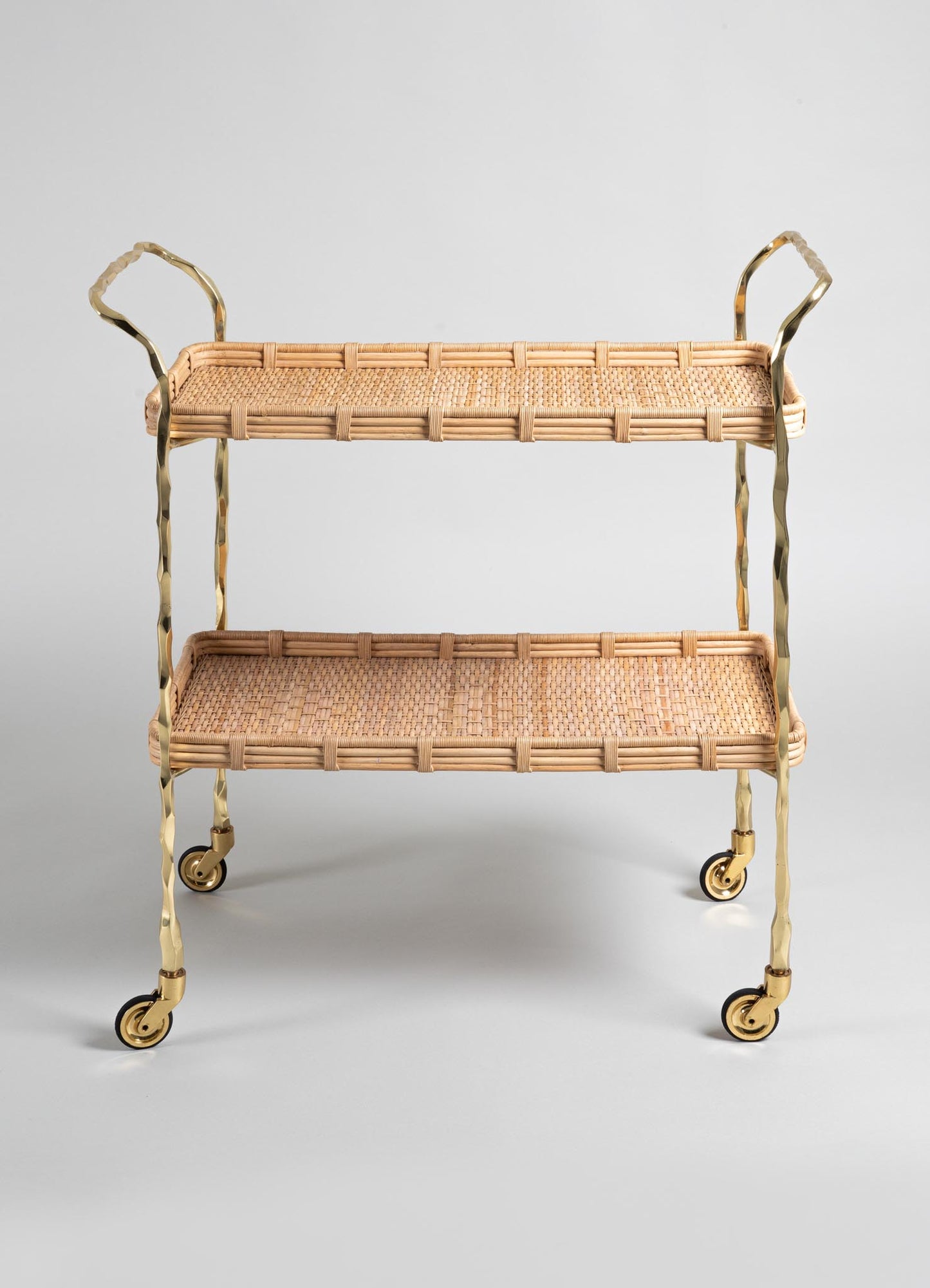 The VIYA Assam Brass & Cane Trolley features two tiers, forged brass rod legs and handles, four wheels, and a woven cane wicker design in a light natural finish, all set against a plain gray background.