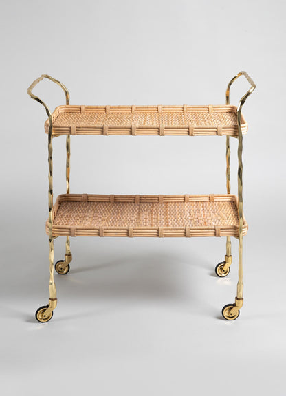 The VIYA Assam Brass & Cane Trolley features two tiers, forged brass rod legs and handles, four wheels, and a woven cane wicker design in a light natural finish, all set against a plain gray background.