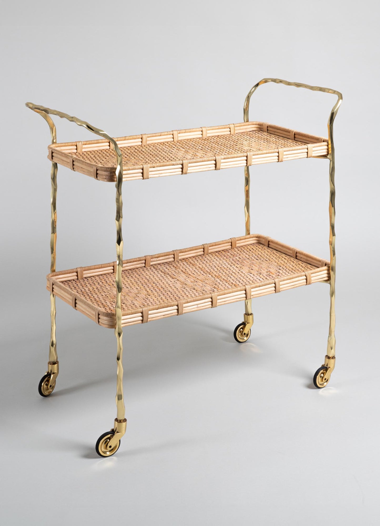 The VIYA Assam Brass & Cane Trolley is a two-tier bar cart with a woven cane design, gold metal frame, curved handles, and four wheels, shown against a light gray background.