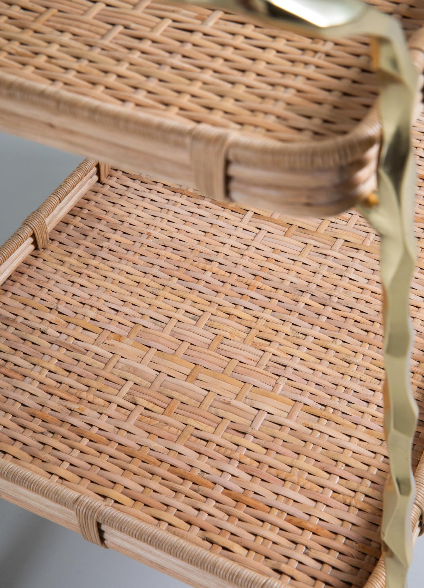 Close-up of two woven cane shelves on the VIYA Assam Brass & Cane Trolley, showing a light natural finish and a partially visible wavy gold handle crafted from forged brass rods on the right.