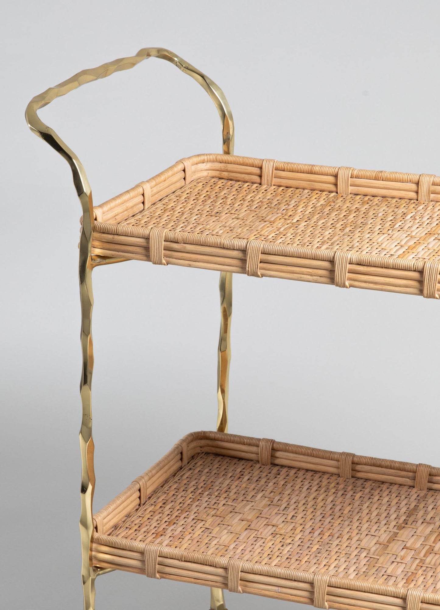 A close-up of VIYA's Assam Brass & Cane Trolley—a two-tiered cart with woven rattan and cane trays, accented by wavy forged brass handles—set against a plain light gray background.
