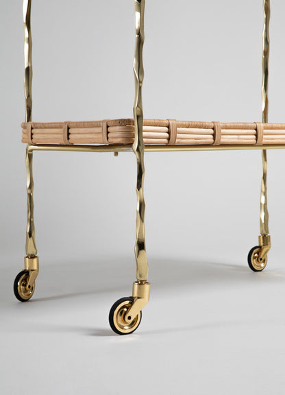A close-up of the Assam Brass & Cane Trolley by VIYA highlights gold textured forged brass rod legs, four small caster wheels, and a woven cane tray shelf against a plain light background.