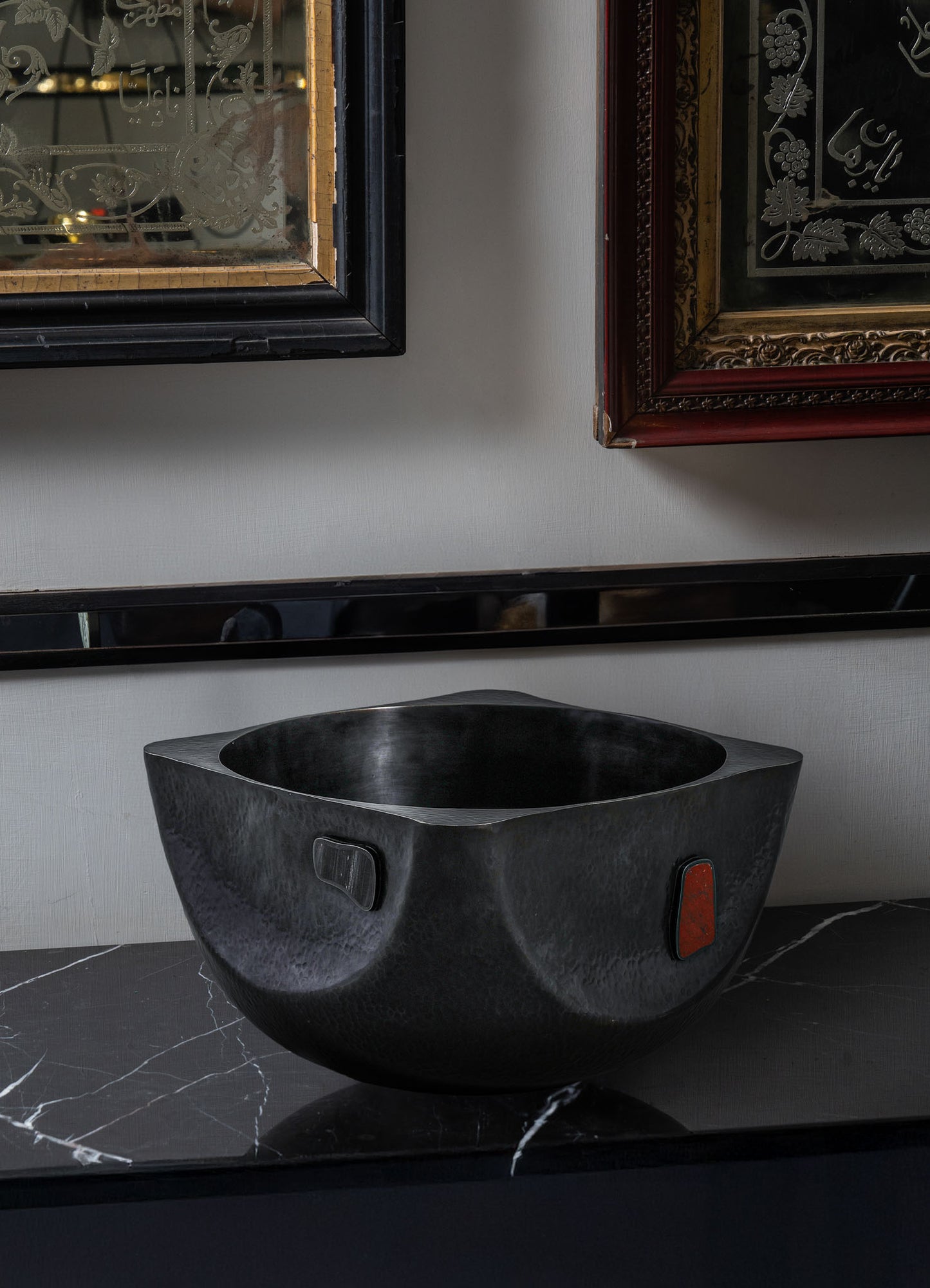 A modern, black, square-shaped Alta Champagne Bucket by VIYA sits on a black marble countertop. Above it, two framed artworks with ornate designs inspired by Alta red dye traditions from the Indian subcontinent hang on the wall.