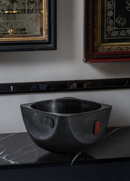 A modern, black, square-shaped Alta Champagne Bucket by VIYA sits on a black marble countertop. Above it, two framed artworks with ornate designs inspired by Alta red dye traditions from the Indian subcontinent hang on the wall.