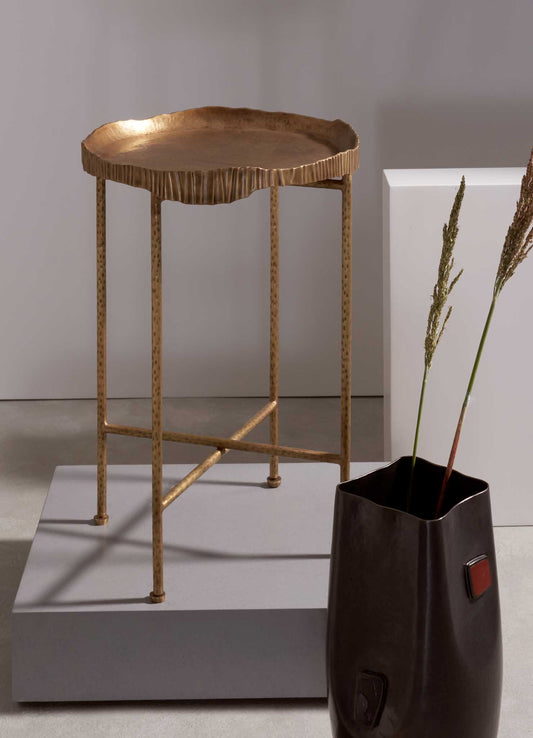 A brass side table with a hammered, irregular-edge top and slender, perforated legs, finished in a warm, golden tone, placed on a neutral surface. A dark ceramic vase with a square shape and an organic, slightly wavy rim holds dried grasses, creating a minimalist and contemporary aesthetic.
