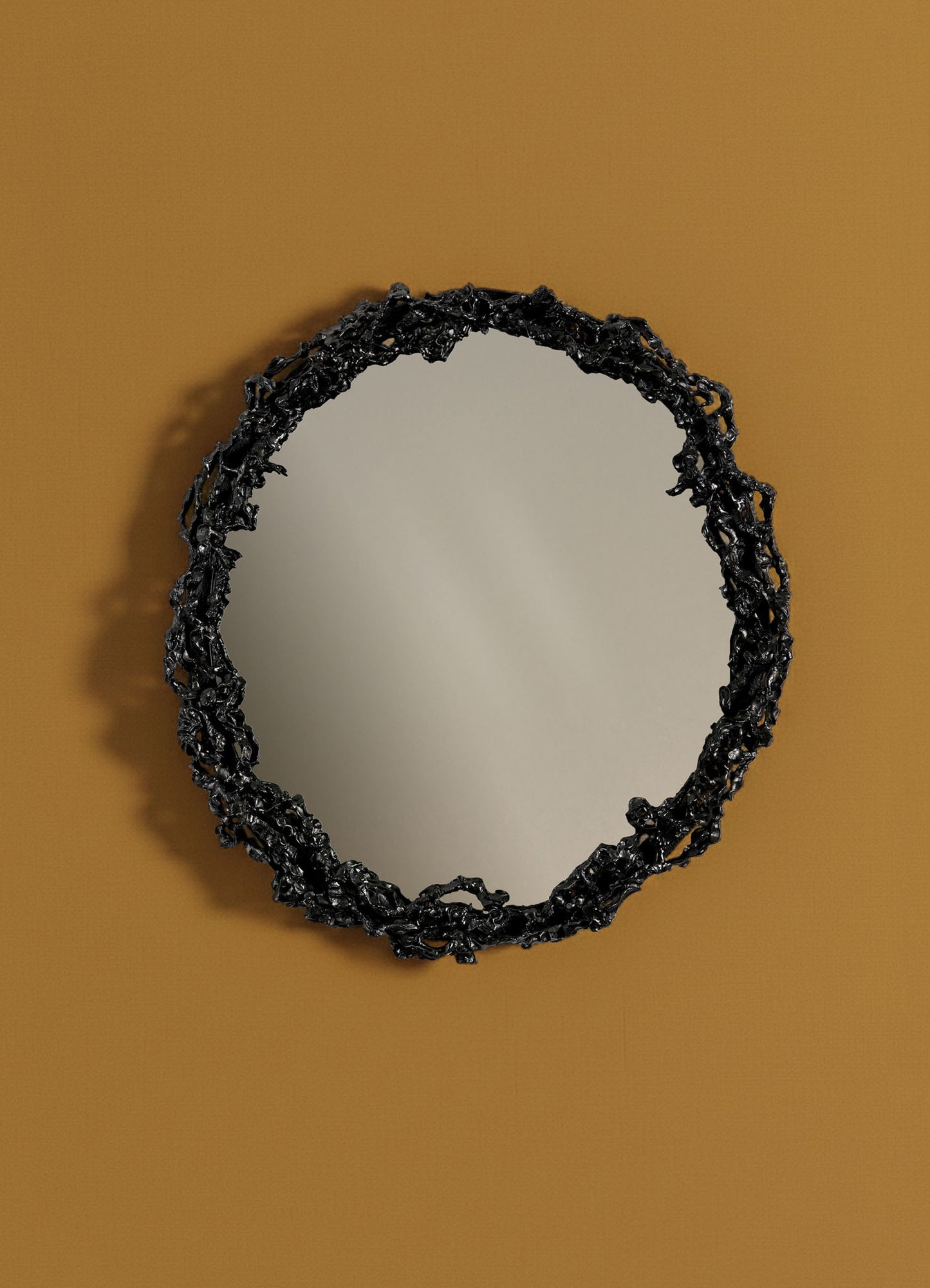 The VIYA Andaman Mirror Round, featuring an ornate textured black frame, hangs on a mustard yellow wall and reflects soft light.