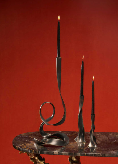 Three tall black candles in modern VIYA Blackbuck Candle Holder Large stand on a dark marble table against a red background. The candle flames are lit.