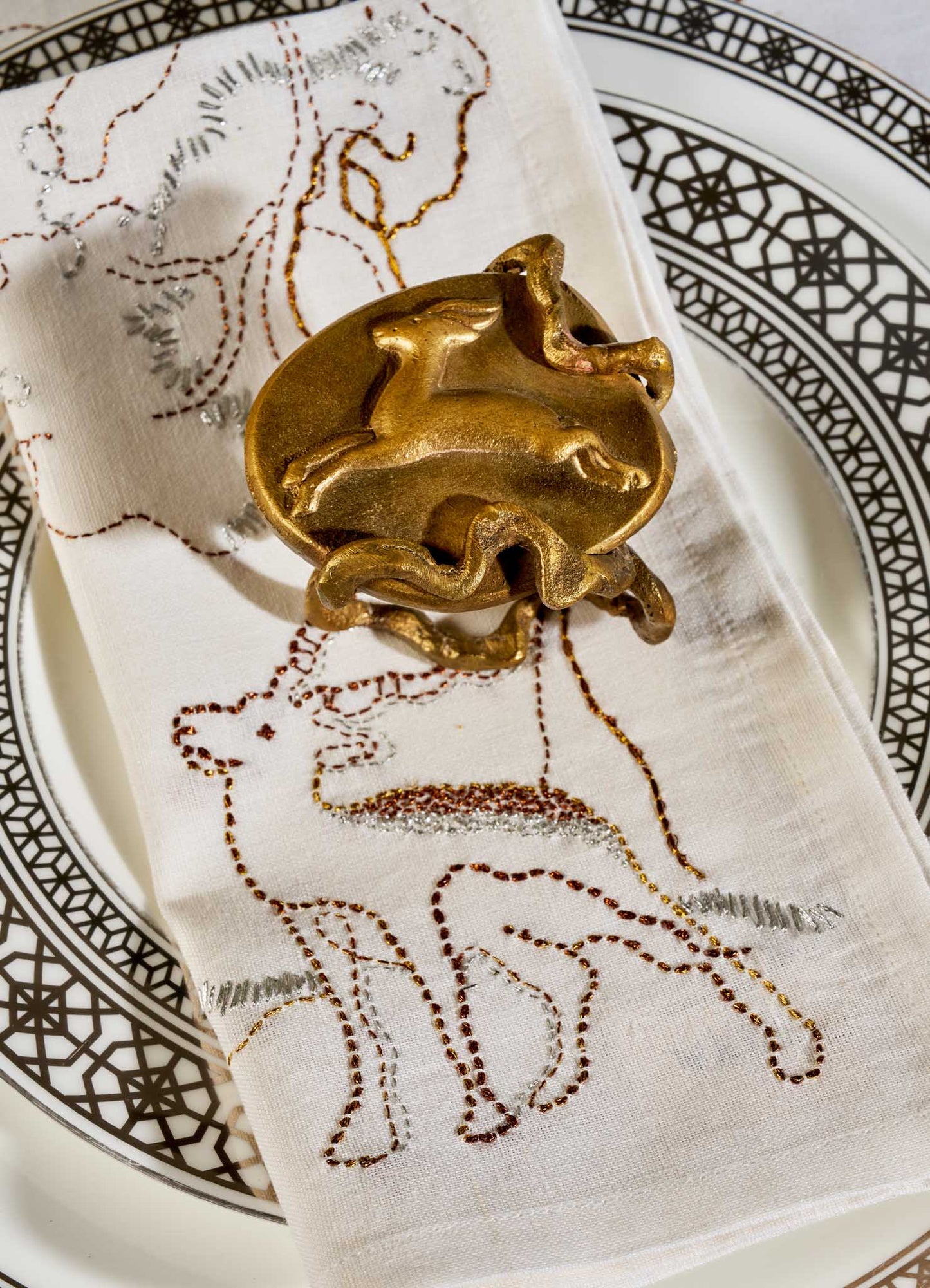 A Viya Blackbuck Dreams Hand Embroidered White & Gold Napkin with animal motifs sits on a decorative plate, secured by a gold napkin ring with abstract animal shapes.
