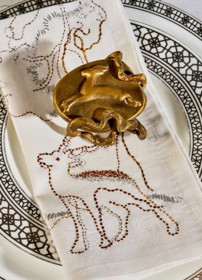 A Viya Blackbuck Dreams Hand Embroidered White & Gold Napkin with animal motifs sits on a decorative plate, secured by a gold napkin ring with abstract animal shapes.