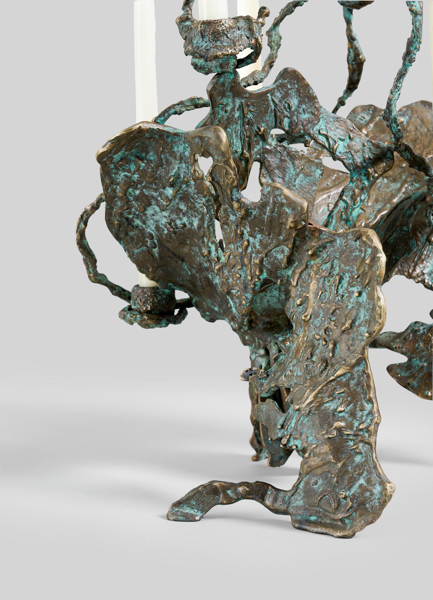 A close-up of the VIYA Butterfly Candle Stand Large shows its abstract bronze form with a rough, greenish-blue patina and twisting shapes. Two white candle tops are visible at the upper edge.