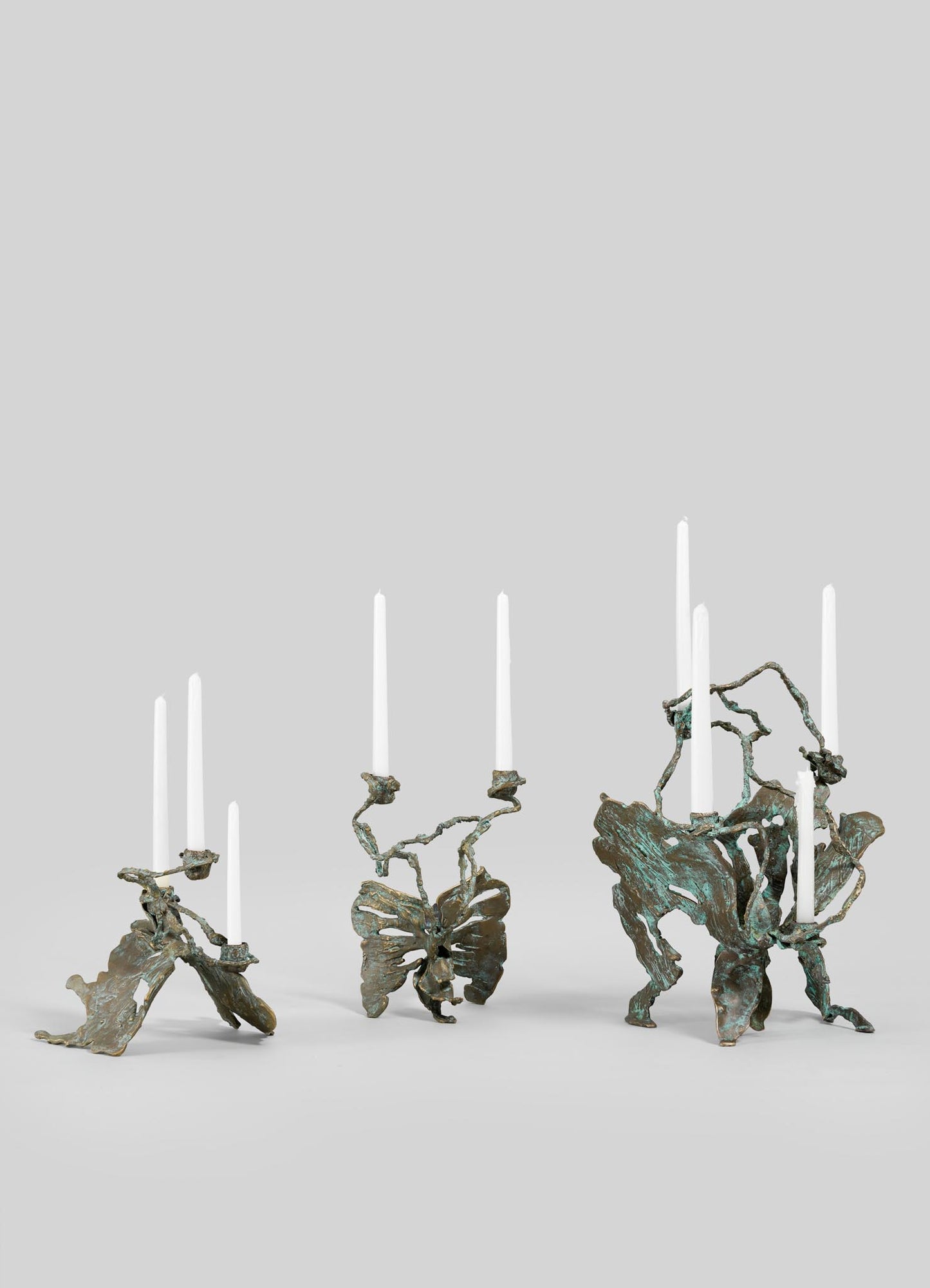 Three VIYA Butterfly Candle Stand Small holders, sculptural bronze pieces with abstract, organic forms, each display tall white taper candles against a plain light gray background.