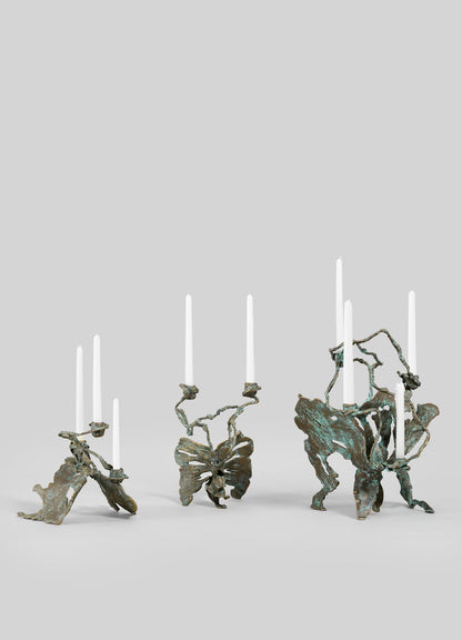 Three VIYA Butterfly Candle Stand Small holders, sculptural bronze pieces with abstract, organic forms, each display tall white taper candles against a plain light gray background.