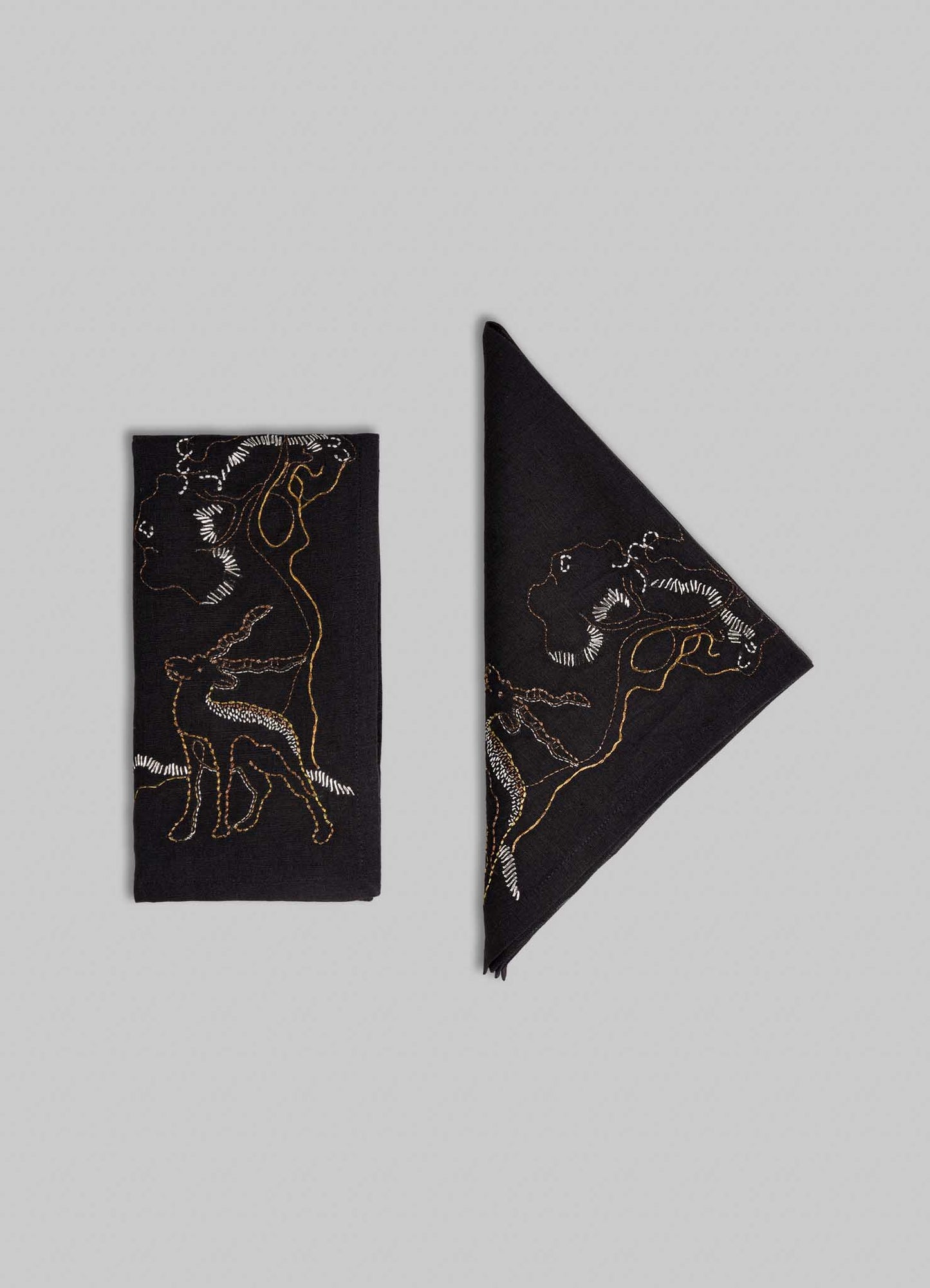 Two Viya Blackbuck Dreams Hand Embroidered Black & Gold Napkins, one rectangular and one diagonal, display abstract human and animal line art, including a Blackbuck, in white and gold thread on black fabric against a light gray background.