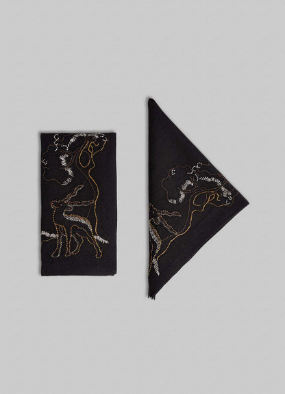 Two Viya Blackbuck Dreams Hand Embroidered Black & Gold Napkins, one rectangular and one diagonal, display abstract human and animal line art, including a Blackbuck, in white and gold thread on black fabric against a light gray background.