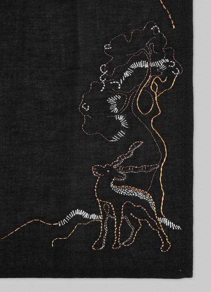 The Viya Blackbuck Dreams Hand Embroidered Black & Gold Napkin features colorful outlines of a woman with a vessel, a deer, and a tree on black fabric, stitched in white, orange, and yellow for an Indian manuscript-inspired look.