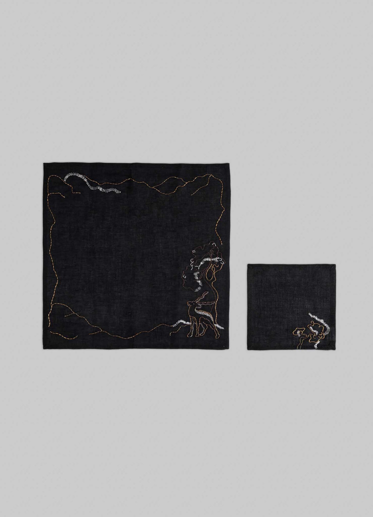 The Viya Blackbuck Dreams Hand Embroidered Black & Gold Napkin set features two black napkins—one larger with a figure smoking amid abstract gold accents, and a smaller piece with an Indian manuscript-inspired motif.