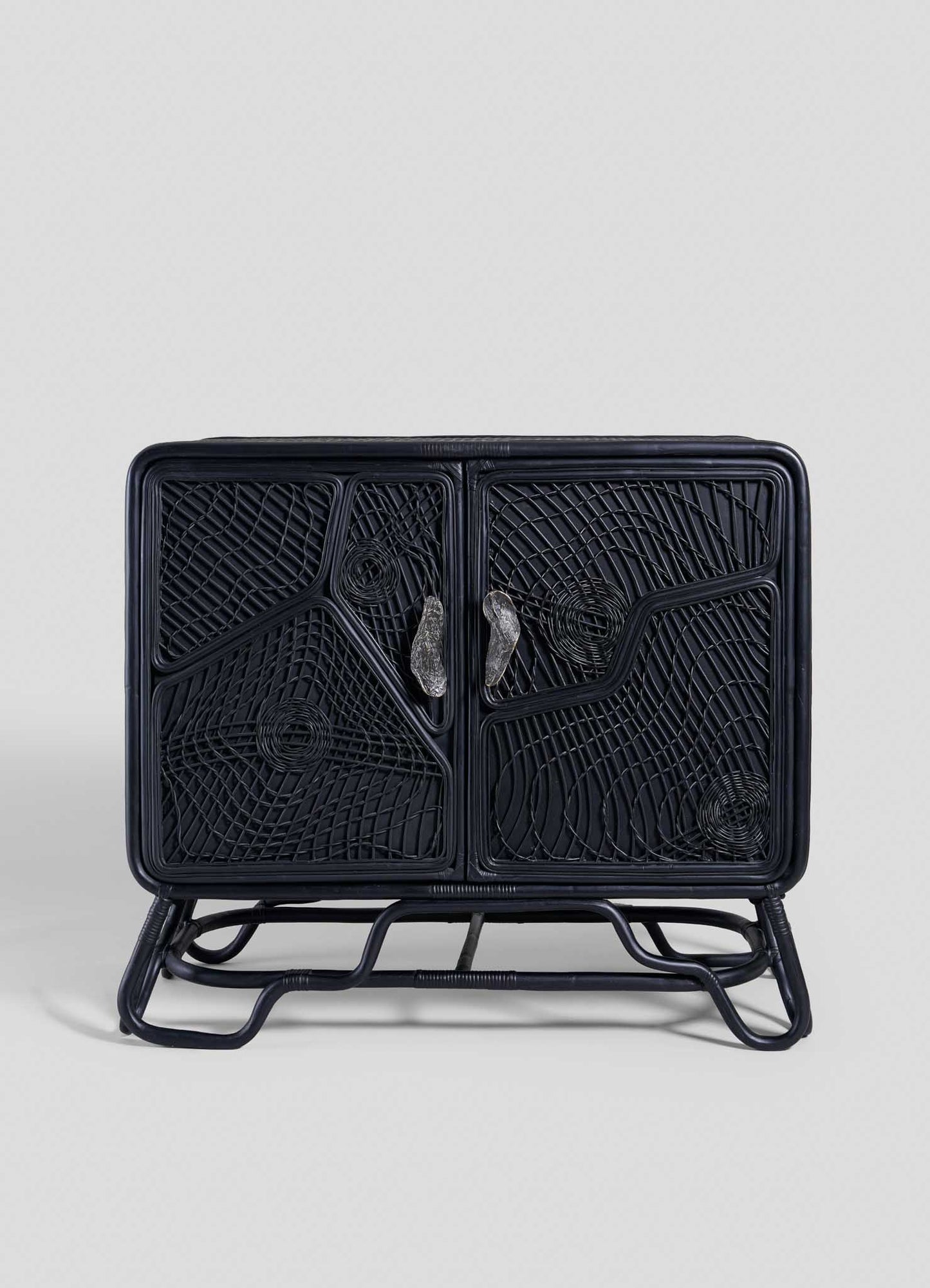 The VIYA Chakra Cabinet features two front doors with geometric cane weaving, curved legs, and two clear handles, all in black rattan, set against a plain light gray background.