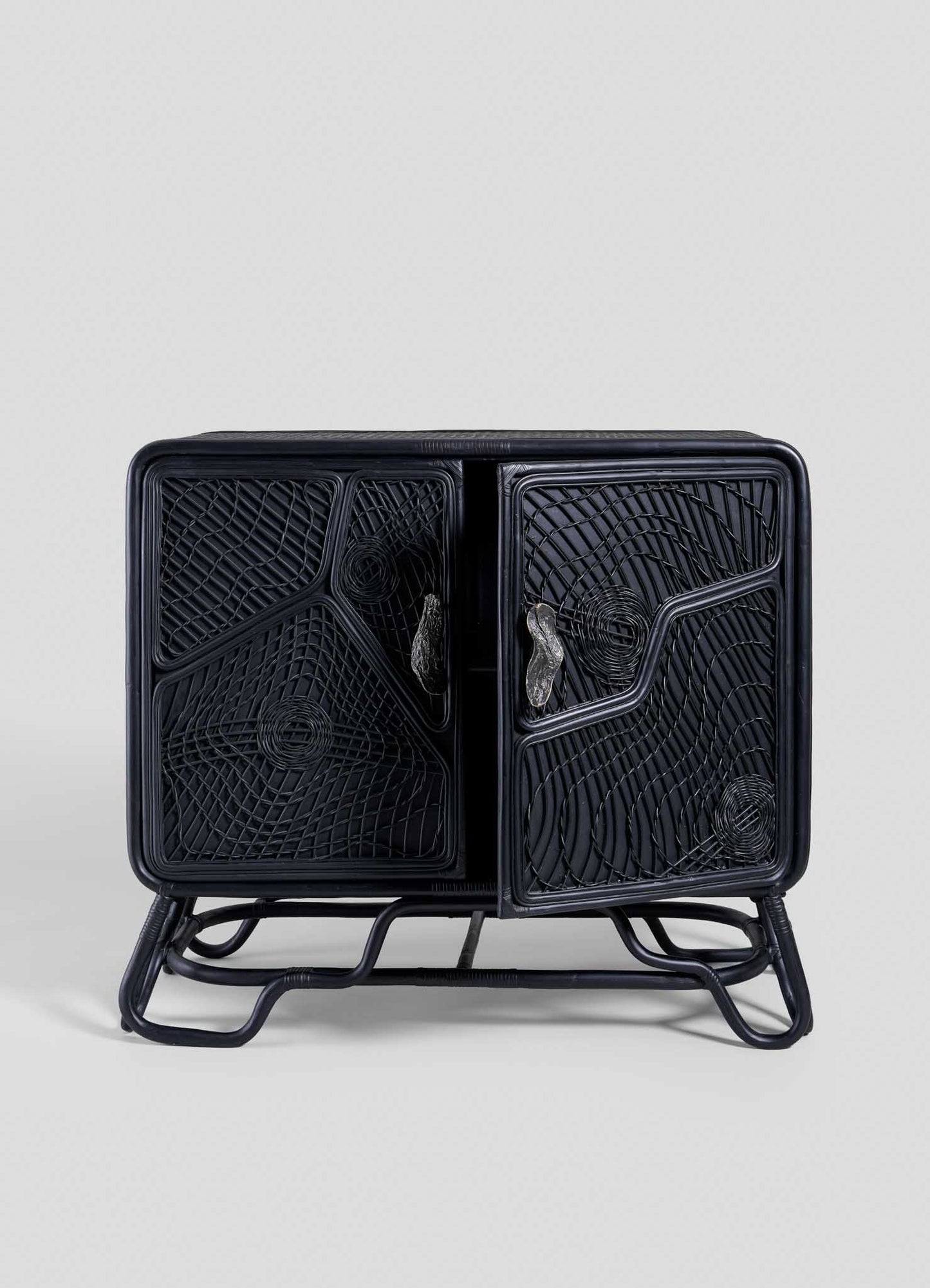 The VIYA Chakra Cabinet is a black rattan cabinet with two doors and geometric cane patterns, set on a matching frame, shown against a plain light gray background.