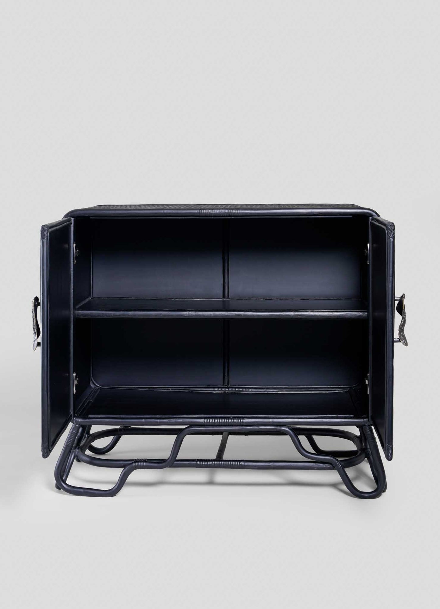 The VIYA Chakra Cabinet is a black metal industrial-style piece with two doors, one interior shelf, and sturdy tubular legs. Shown empty against a plain light background, it’s perfect to pair with rattan plants for stylish storage.