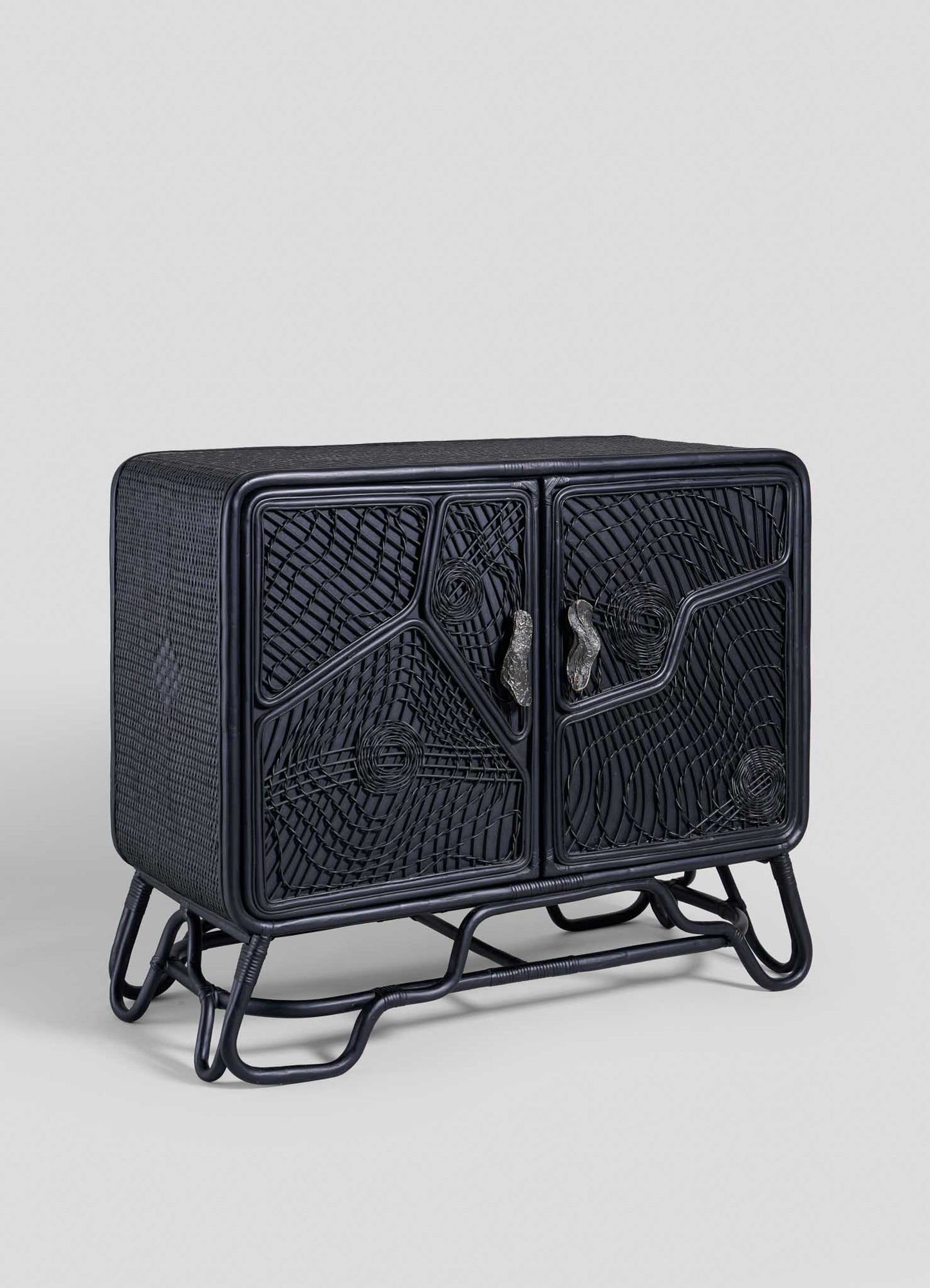 The Chakra Cabinet by VIYA features intricate cane-weaved black doors and a tubular metal base, presented against a plain light gray background.