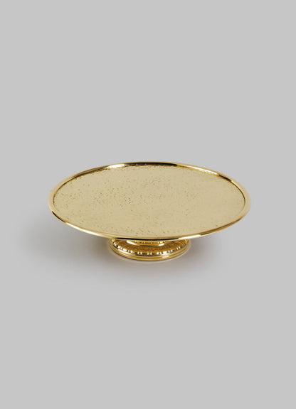 The VIYA City Palace Cake Platter is a round, gold-colored handcrafted brass stand with a textured surface and short decorative pedestal base, shown against a plain light gray background.