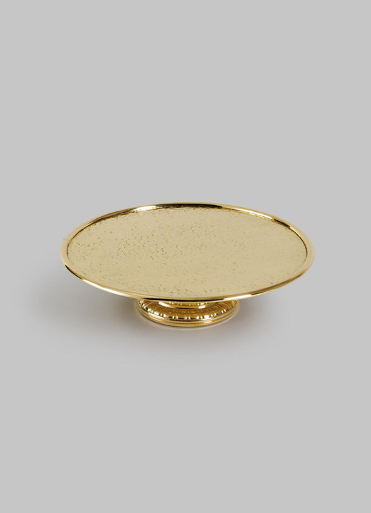 The VIYA City Palace Cake Platter is a round, gold-colored handcrafted brass stand with a textured surface and short decorative pedestal base, shown against a plain light gray background.