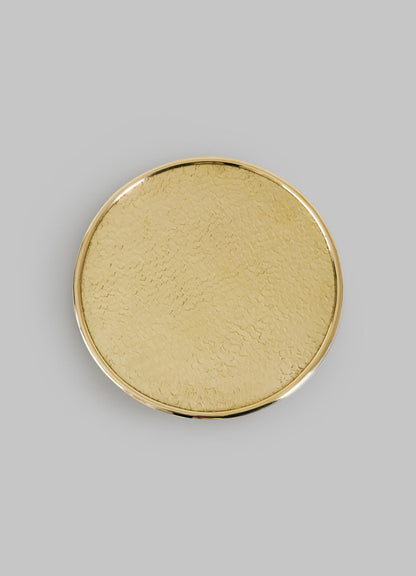 The VIYA City Palace Cake Platter is a round, gold-toned handcrafted brass tray with a hammered texture and smooth raised edge, inspired by Jaipur in Bloom, shown on a plain light gray background.