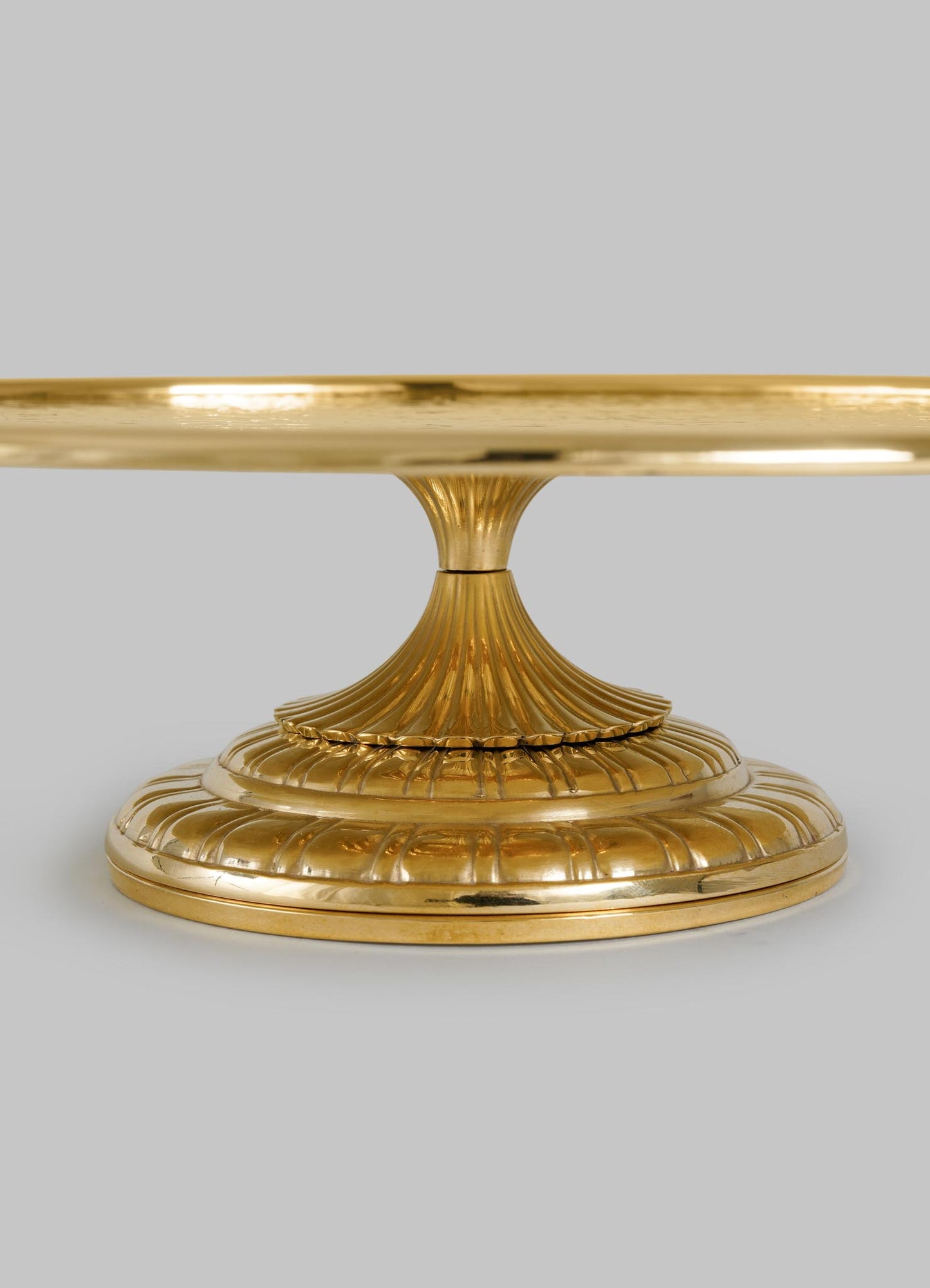 The VIYA City Palace Cake Platter is a gold pedestal stand inspired by Jaipur in Bloom, featuring a fluted stem and ribbed base. This handcrafted brass piece stands elegantly against a plain gray background.