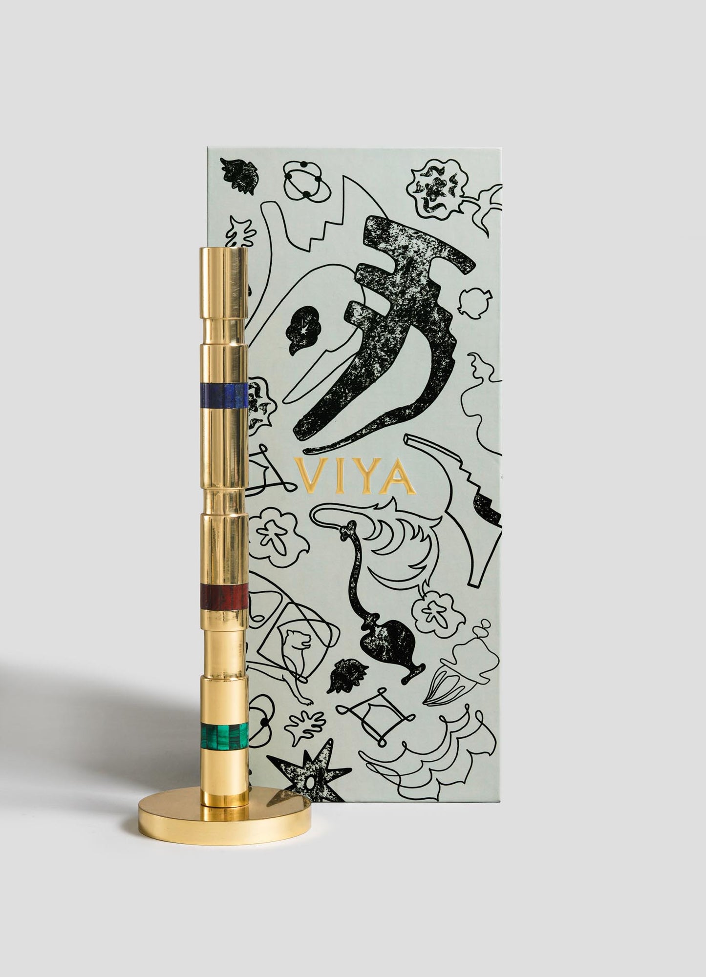The Eros Candle Holder by VIYA features a gold-toned cylindrical design with colored bands and is displayed before an Art Deco-inspired box adorned with abstract black illustrations and "VIYA" in gold, echoing the glamour of vintage Bombay cinema.