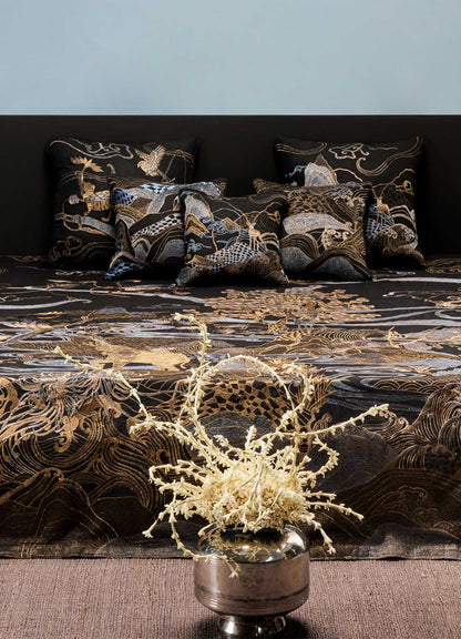 A Viya Dreamscape Black & Gold Embroidered Bedcover decorates the bed with cushions of symbolic motifs, set against a plain light blue wall. A silver vase filled with dried beige branches stands in the foreground.