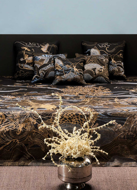 A Viya Dreamscape Black & Gold Embroidered Bedcover decorates the bed with cushions of symbolic motifs, set against a plain light blue wall. A silver vase filled with dried beige branches stands in the foreground.