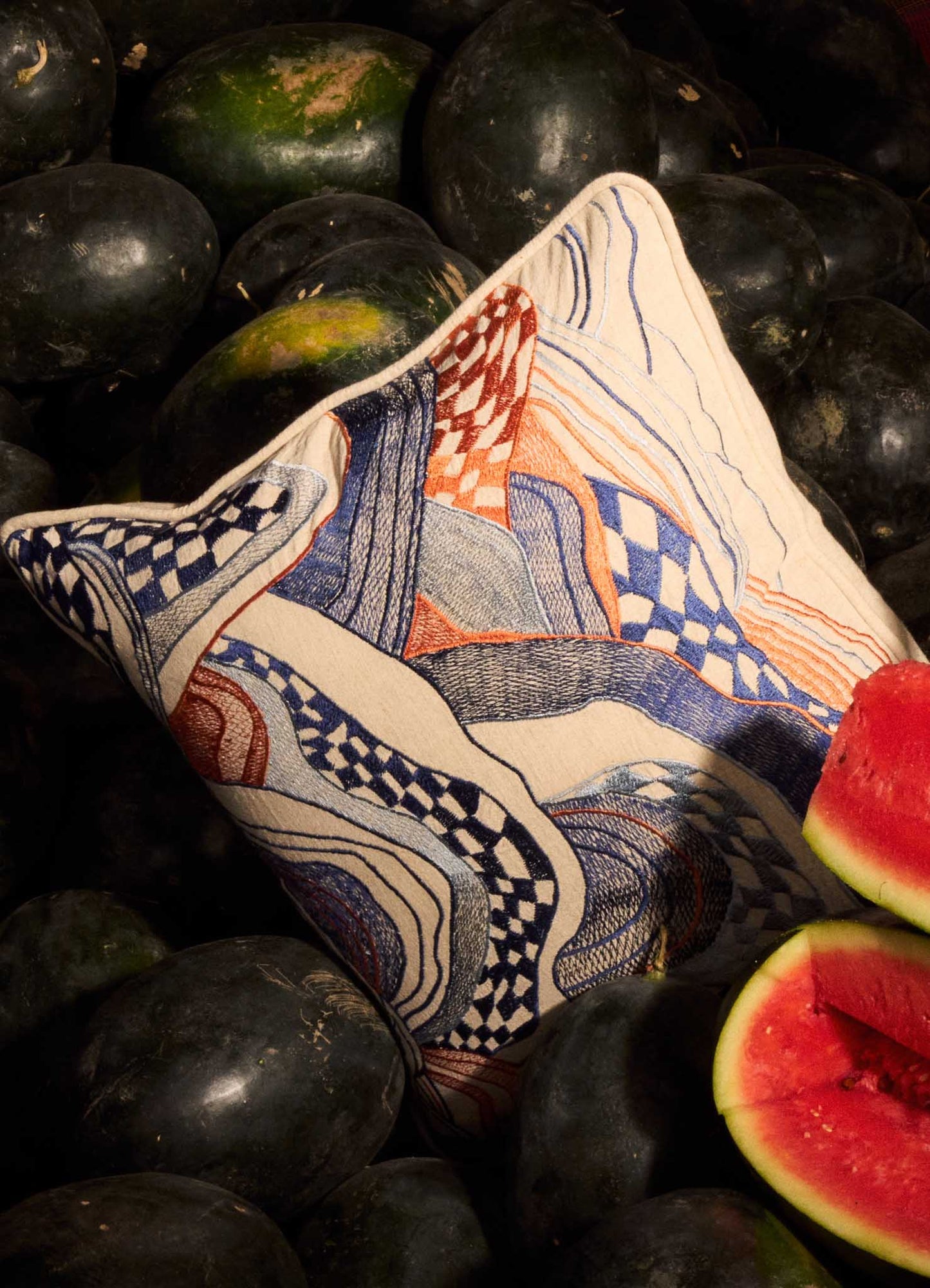 The Viya Dreamscape Blue Embroidered Cushion Cover 18"x18" with abstract red, blue, and orange motifs sits among dark green watermelons, one sliced open to reveal its bright red flesh in the foreground.