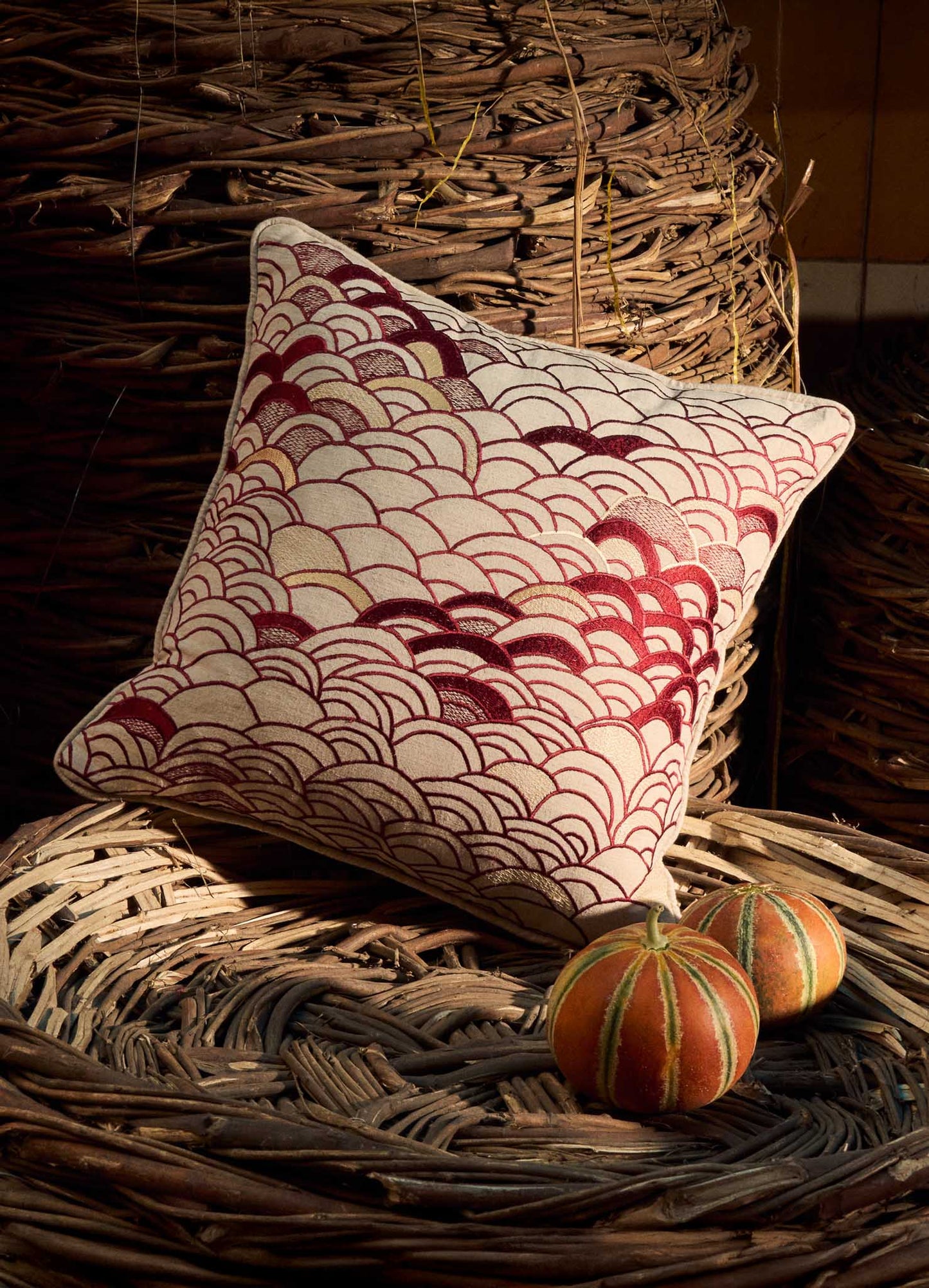 The Viya Dreamscape Red Embroidered Cushion Cover B (18"x18") with a red and white scallop pattern rests on a woven basket, while two small orange-and-green gourds nearby are illuminated by sunlight.