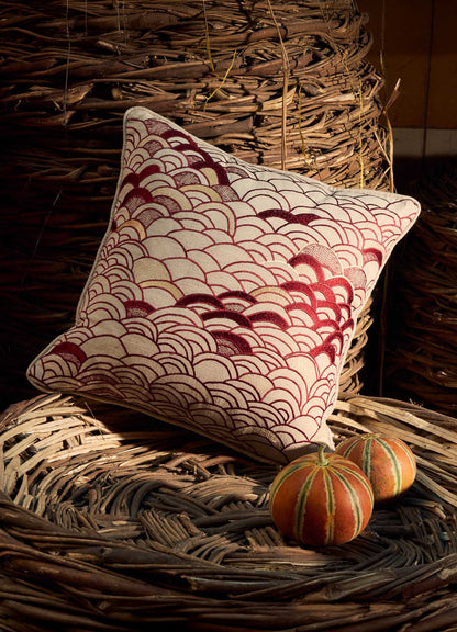 The Viya Dreamscape Red Embroidered Cushion Cover B (18"x18") with a red and white scallop pattern rests on a woven basket, while two small orange-and-green gourds nearby are illuminated by sunlight.