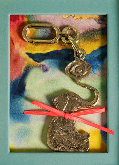 The Eye Of The Elephant Charm by VIYA is a gold elephant keychain with an abstract evil eye above its trunk, accented by a pink ribbon bow for added protection and charm, displayed on a colorful watercolor background.