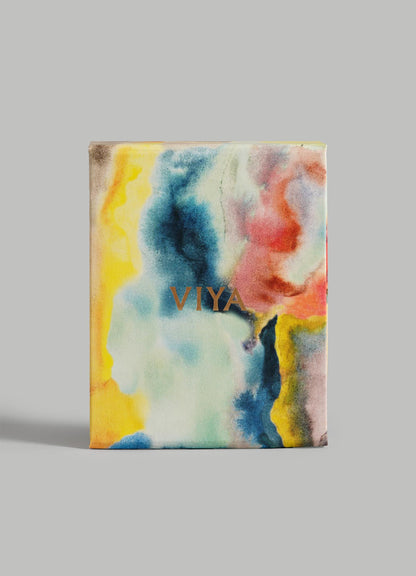 The Eye Of The Elephant Charm by VIYA is a square piece with an abstract watercolor design in yellow, blue, pink, and black tones. "VIYA" appears in gold at the center, evoking evil eye protection, shown here against a plain gray background.