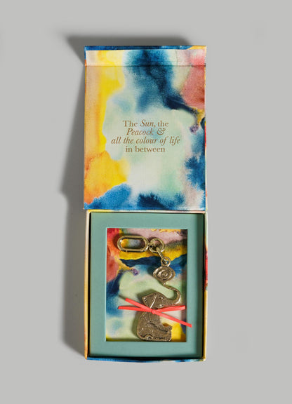 A vibrant VIYA box with abstract watercolor art opens to reveal The Eye Of The Elephant Charm keychain, decorated with brass charms for protection. Inside the lid: The Sun, the Peacock & all the colour of life in between.