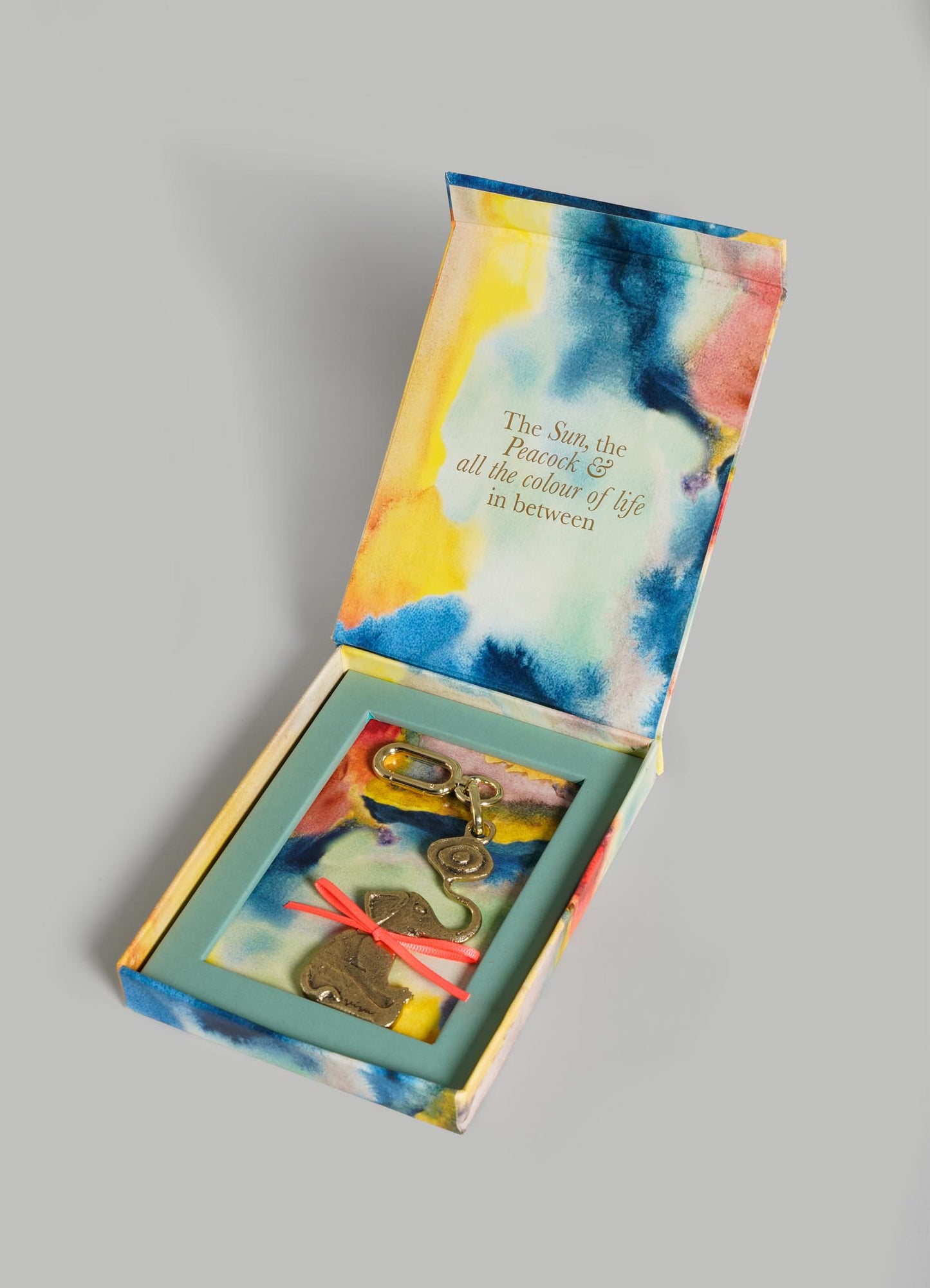 An open, colorful tie-dye box from VIYA reveals The Eye Of The Elephant Charm keychain with brass charms and abstract shapes. Inside the lid: "The Sun, the Peacock & all the colour of life in between—symbols of protection.