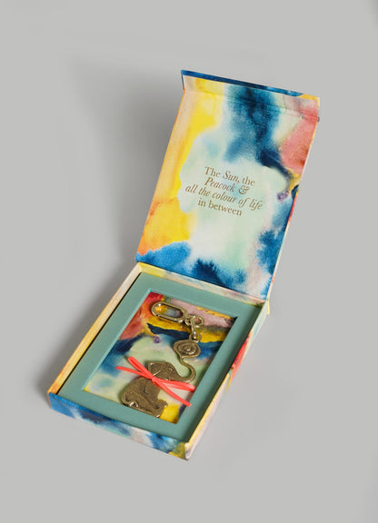 An open, colorful tie-dye box from VIYA reveals The Eye Of The Elephant Charm keychain with brass charms and abstract shapes. Inside the lid: "The Sun, the Peacock & all the colour of life in between—symbols of protection.