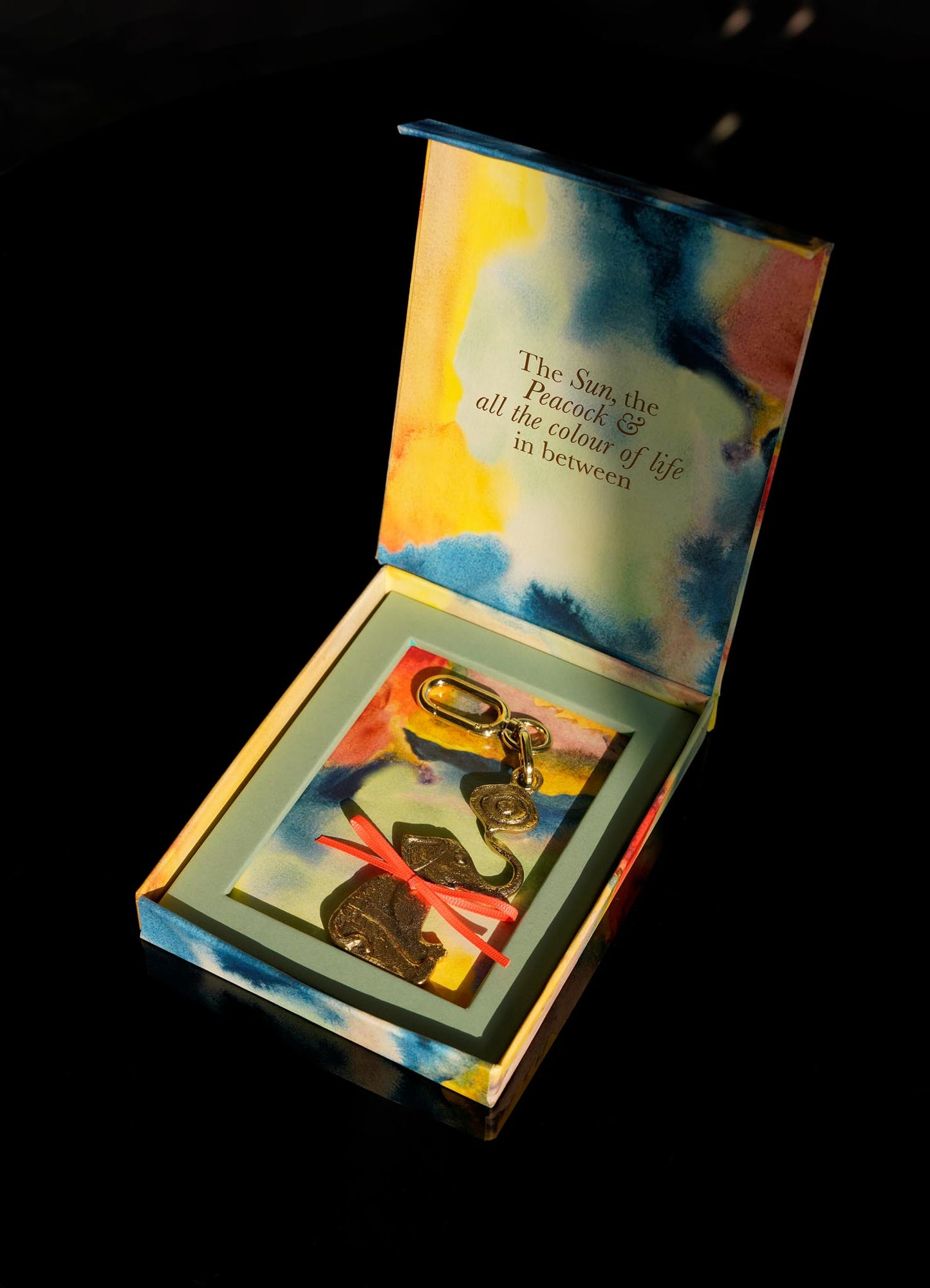 An open, colorful VIYA box with abstract patterns reveals The Eye Of The Elephant Charm—a gold keychain with evil eye and brass charms tied by a pink ribbon. Inside the lid: "The Sun, the Peacock & all the color of life in between.