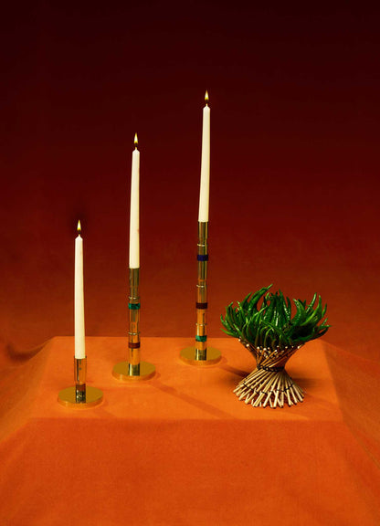 Three white taper candles in VIYA Liberty Candle Holders are arranged on an orange surface beside a small green plant, evoking Art Deco elegance against a striking red-orange gradient background.
