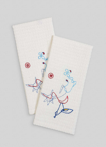 The Viya Evil Eye Hand Embroidered Ecru Hand Towels (Set of 2) feature abstract, colorful Nazar-inspired designs in blue, red, and black depicting flowers and stars on a plain gray background.