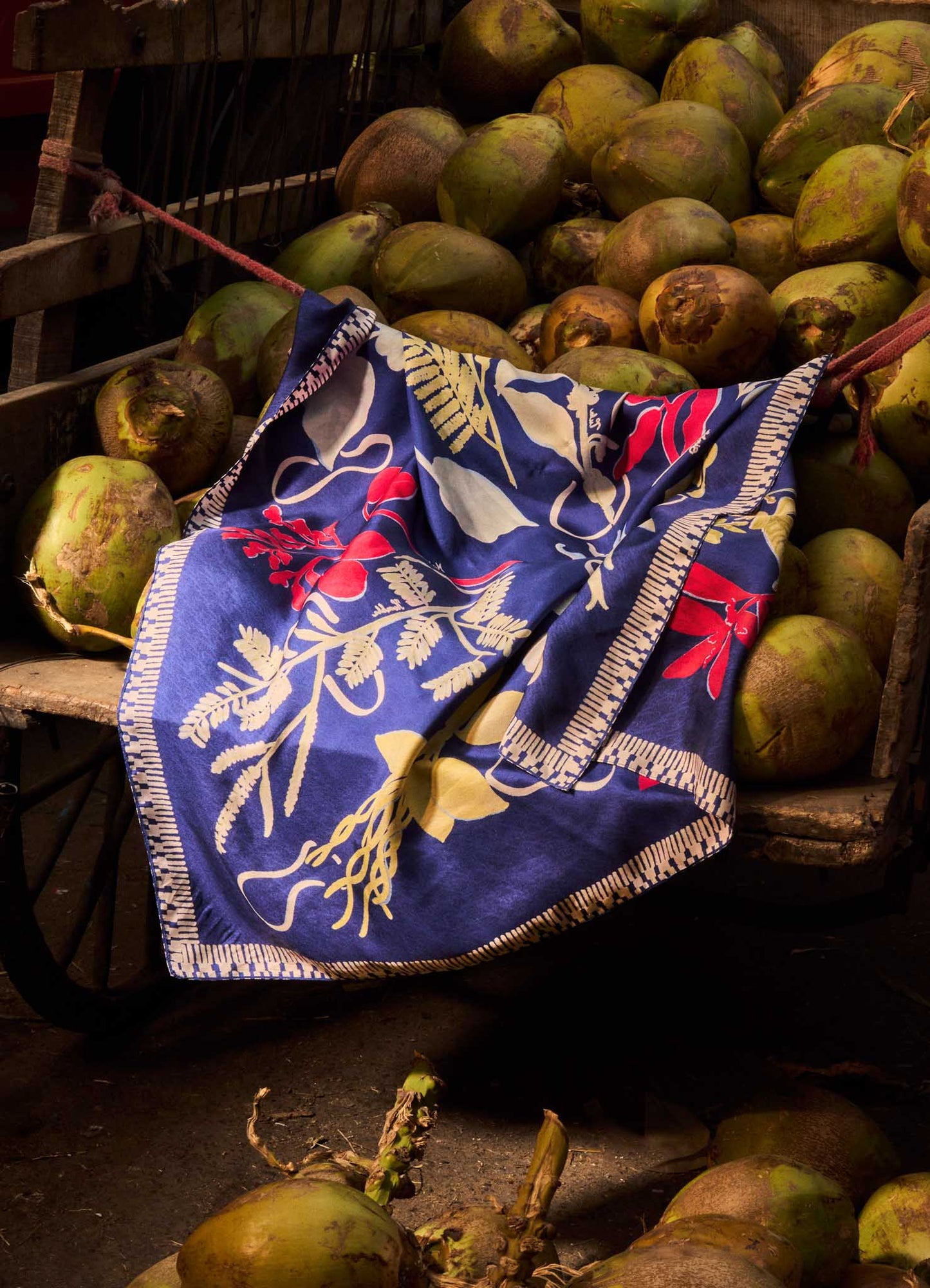 The Viya Floral Astra Navy Silk Scarf, featuring red, yellow, and white Indian floral motifs, is draped over green coconuts in a rustic cart.